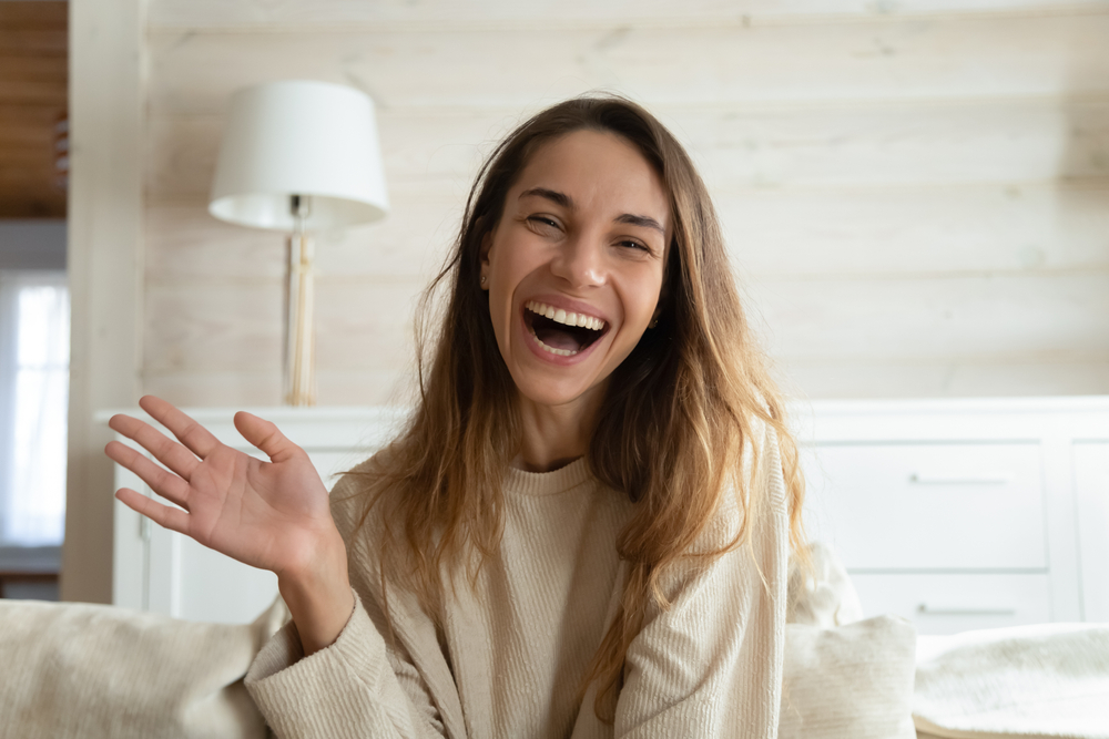 Smiling young woman waving happily at home, showcasing confidence restored by Dental Crowns at West Kerr Dental